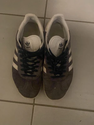 Adidas Gazelle grå sneakers - Säljer ett par klassiska Adidas Gazelle sneakers i grå mocka med vita detaljer och svarta skosnören. Skorna har de ikoniska tre vita ränderna på sidan och vit plös med Adidas-logga. Perfekta för dig som gillar retrostil och streetwear.