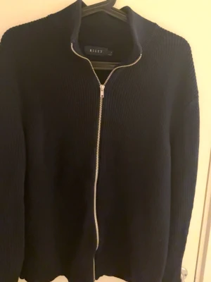 Svart stickad ziptröja från Riley - Säljer en svart stickad tröja från Riley med hel dragkedja framtill och hög krage. Tröjan har lång ärm och är ribbstickad för en snygg och enkel look. Perfekt för lager-på-lager och passar till det mesta.