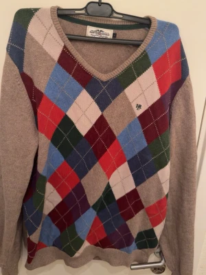 Beige argyle stickad tröja RL - Stickad tröja från  Lauren med klassiskt argylemönster i beige, blått, rött, grönt, vitt Rl vinrött framtill. V-ringad hals och långa ärmar. Materialet känns mjukt och tröjan har RL-logga broderad på bröstet. Perfekt för en preppy look.