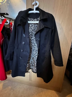 Svart trenchcoat från Divided - Stilren svart trenchcoat från Divided med dubbelknäppning och klassisk krage. Insidan har ett coolt leopardmönstrat foder som ger jackan en unik touch. Perfekt för dig som vill ha en trendig och tidlös look. Materialet känns lätt och smidigt.