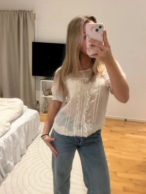 Vit romantisk blus med spetsdetaljer - Söt vit blus i transparent material med korta puffärmar och dekorativa spetsdetaljer längs bröstet och ärmsluten. Blusen har volangkant framtill och en feminin, luftig passform. Perfekt för en trendig och romantisk look.passar s-l