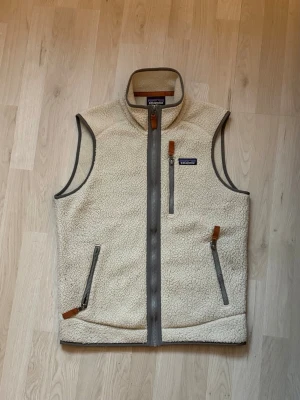 Beige fleeceväst från Patagonia - Säljer en beige fleeceväst från Patagonia med grå detaljer och dragkedjor. Västen har hög krage, tre fickor med dragkedja och snygga läderdetaljer på dragkedjorna. Perfekt för lager-på-lager och chill stil.