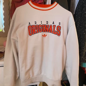Krämvit Adidas Originals sweatshirt - Snygg vit sweatshirt från Adidas Originals med broderad logga och text i svart och rött på bröstet. Tröjan har ribbade muddar och krage med orange och vita ränder. Perfekt för en sportig och avslappnad stil. Storlek oversize XS men mer som en S/M. 