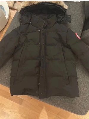 Svart dunjacka från Canada Goose Wyndham - Säljer en svart dunjacka från Canada Goose, modell Expedition Parka. Jackan har huva med fuskpäls, stora fickor och klassisk logga på ärmen. Perfekt för kalla vinterdagar med sin tjocka vaddering och vindtäta material.
