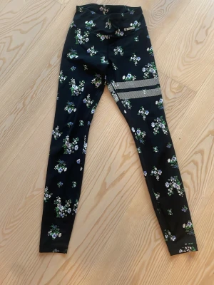 Blommiga leggings från Stronger - Snygga svarta leggings från Stronger med blommigt mönster i vitt, grönt och lila. Hög midja och dekorativa ränder på ena benet. Tillverkade i stretchigt funktionsmaterial som passar perfekt till träning eller chill.