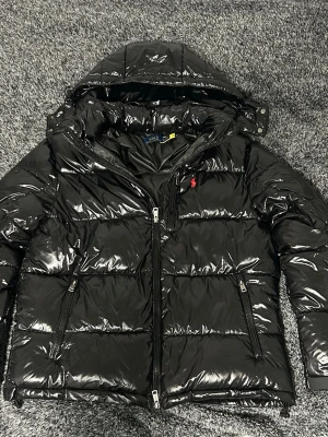 Svart glansig dunjacka Polo Ralph Lauren - Säljer en svart, glansig dunjacka från Polo Ralph Lauren med huva och dragkedja framtill. Jackan har två sidofickor med dragkedja och den klassiska röda loggan på bröstet. Perfekt för kalla vinterdagar och riktigt snygg streetstil.