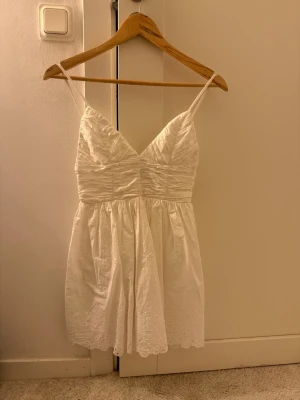 Vit kort sommarklänning/studentklänning - Söt vit kortklänning med smala axelband och v-ringning. Klänningen har rynkade detaljer upptill och spetsmönster nertill på kjolen. Perfekt för varma dagar och har en luftig, lätt känsla.