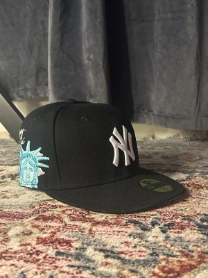 Svart New Era Yankees keps - Svart keps från New Era med New York Yankees-logga i vitt framtill och broderad Frihetsgudinna i blått och vitt på sidan. Klassisk flatbrätte och broderad New Era-logga på sidan. Perfekt för dig som gillar streetwear och sportig stil.