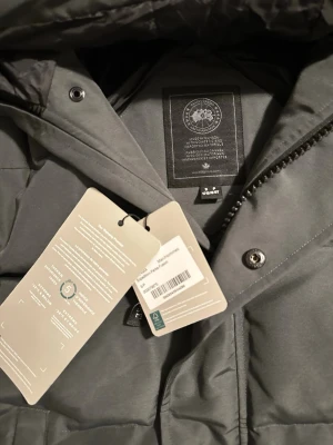 Grå dunjacka från Canada Goose - Snygg grå dunjacka från Canada Goose med huva och dragkedja. Jackan har två stora framfickor, diskret logga på ärmen och stilren design. Perfekt för kalla vinterdagar och håller dig varm med sin tjocka vaddering.