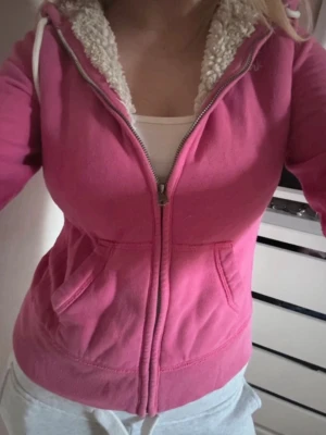 Rosa hoodie från Abercrombie & Fitch - Säljer en rosa hoodie från Abercrombie & Fitch med dragkedja och två fickor framtill. Insidan av huvan är fodrad med mjuk, vit teddy. Broderad logga 'A&F New York' på bröstet. Perfekt för en avslappnad och trendig look.