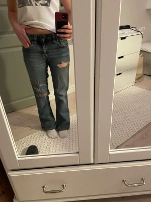 Blå bootcut jeans med slitningar - Säljer ett par blå bootcut jeans med snygga slitningar på båda benen. Jeansen har klassisk femficksdesign och låg midja. Perfekta för en avslappnad och trendig look.