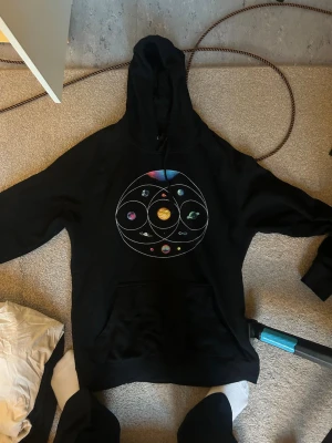 Coldplay Music of the Spheres hoodie - Svart hoodie med färgglatt planetsystem-tryck på bröstet och texten 'COLDPLAY' på ryggen. På ena ärmen står det 'MUSIC of the SPHERES'. 