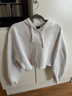 Vit cropped hoodie med huva - Säljer en vit cropped hoodie från Bik Bok med huva och dragsko. Hoodien har långa ärmar med ribbade muddar och är gjord i mjukt sweatshirtmaterial. Perfekt för en avslappnad och trendig look.
