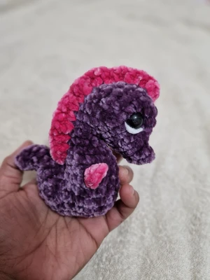 Lila och rosa virkad sjöhäst - En söt, handgjord sjöhäst i mjuk lila och rosa garn. Den har stora svarta ögon och en rosa man längs ryggen. Perfekt som dekoration eller liten maskot. Tillverkad i plyschliknande material.