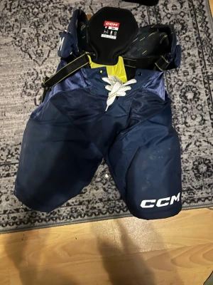 Blå hockeybyxor från CCM - Säljer ett par blå hockeybyxor från CCM med vita detaljer och snörning framtill. Byxorna har vaddering för extra skydd och är tillverkade i slitstarkt syntetmaterial. Perfekta för dig som spelar ishockey och vill ha bra skydd på isen.