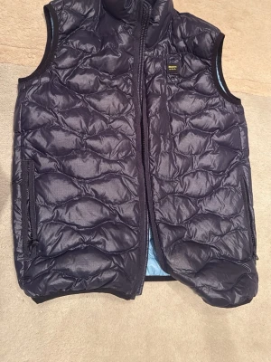 Skjut snygg blauer mörkblå väst - Snygg svart dunväst med quiltat mönster och hög krage. Västen har två dragkedjeförsedda fickor på sidorna och en liten logotyp på bröstet. Insidan är ljusblå och materialet är glansigt och lätt. Perfekt för lager-på-lager och streetwear. 14 år/164, nypris 1600