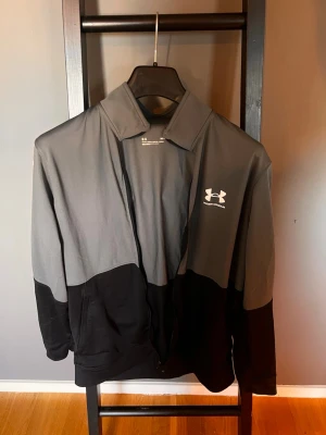 Under Armour grå och svart träningsjacka - Snygg träningsjacka från Under Armour i grått och svart med dragkedja framtill och hög krage. Jackan har vit logga på bröstet och reflekterande detaljer på ryggen. Perfekt för träning eller sportiga dagar. Jätte bra skick men drag kedjan e lite sönder men går nog att lösa lätt därav priset 