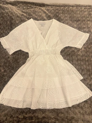 Vit klänning  - Söt vit kortklänning med broderade blommor, omlott-v-ringning och volangdetaljer. Klänningen har korta ärmar och smock i midjan för en bekväm passform. Perfekt för studenten och sommarens alla tillfällen. Klänning har aldrig använts, prislapp sitter kvar. Det står storlek 32 men pga den väldigt stretchiga midjan så passar den alla från storlek 32-36.  