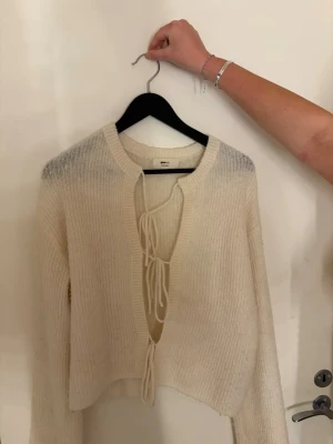 Krämvit stickad cardigan med knyt - Trendig krämvit stickad cardigan från Gina med långa ärmar och två knytband framtill. Croppad modell med bred ribbstickad kant och mjukt material som känns skönt mot huden. Perfekt att styla över ett linne eller topp.