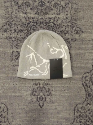 Arc'teryx ljusgrå mössa i ull - Snygg ljusgrå mössa från Arc'teryx med vitt grafiskt mönster och broderad logga framtill. Tillverkad i mjukt stickat material som sitter skönt på huvudet. Perfekt för dig som vill ha en stilren och modern accessoar till vintern.