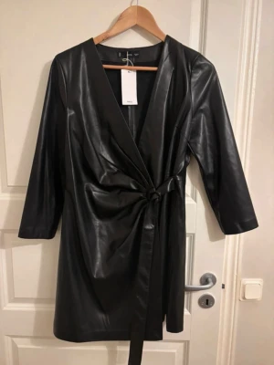 Svart omlottklänning i skinnimitation - Snygg svart omlottklänning från MANGO i skinnimitation med trekvartsärm och knytband i midjan. Klänningen har en v-ringad urringning och kort längd, perfekt för en trendig look. Materialet ger en cool och modern känsla. Kolla min profil för fler plagg! 🥰