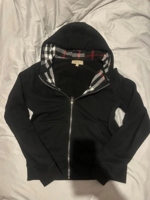 Svart hoodie från Burberry med dragkedja - Snygg svart hoodie från Burberry med klassiskt rutigt foder i huvan och hel dragkedja framtill. Hoodien har långa ärmar och två fickor fram. Perfekt för dig som vill ha en stilren och exklusiv look.