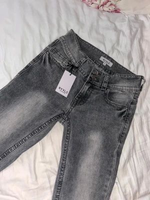 Grå jeans från RYVLS med fickdetaljer - Snygga grå jeans från RYVLS med tvättad look och raka ben. Jeansen har unika bakfickor med lock och knappdetaljer samt klassisk femficksdesign. Perfekta för dig som vill ha en cool och avslappnad stil. Dom är aldrig använda.