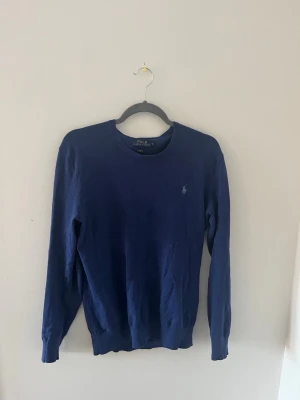 Mörkblå tröja från Polo Ralph Lauren - Klassisk mörkblå tröja från Polo Ralph Lauren med rund halsringning och den ikoniska broderade loggan på bröstet. Tröjan har långa ärmar och ribbade muddar vid ärmslut och nederkant. Perfekt för en stilren och enkel look.