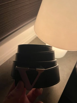 Svart bälte med metallspänne - Snyggt svart bälte med ett stort, triangelformat metallspänne. Bältet har en stilren design med diskreta sömmar längs kanterna och är tillverkat i skinn. Perfekt accessoar för att lyfta din outfit.