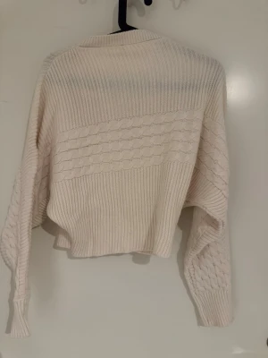 Krämvit stickad croppad tröja - Trendig krämvit stickad croppad tröja från Suwariky med snyggt strukturmönster på både ärmar och över bröstet. Tröjan har långa ärmar och ribbade muddar, perfekt för lager på lager och en chill look.