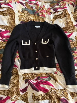 Svart kofta från Holly & Whyte XS - Säljer en svart kofta från Holly & Whyte med vita detaljer och pärlknappar. Koftan har två dekorativa fickor framtill, ribbade muddar och nederkant samt v-ringad hals. Perfekt för dig som gillar klassisk men trendig stil.