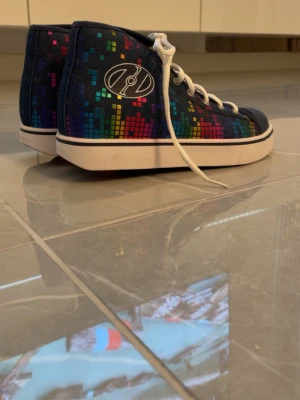 Färgglada sneakers med rullskridskor under sulan - Säljer ett par höga sneakers från JD med svart bas och färgglatt pixelmönster i blått, rött, gult och grönt. Skorna har ett hjul som man kan åka rullskridskor men även är de väldigt snygga. Skorna har vita snören och vit sula samt en rundad tå. Perfekta för dig som vill sticka ut med din stil!