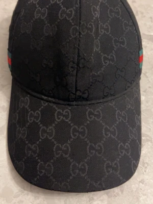 Svart keps från Gucci med monogram - Snygg svart keps från Gucci med klassiskt GG-monogram över hela kepsen. På sidan finns en ikonisk grön och röd rand. Kepsen är tillverkad i ett slitstarkt tygmaterial och har böjd skärm för en clean look.