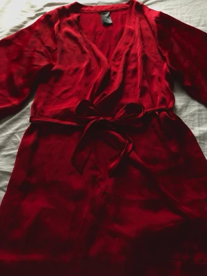 Röd satinmorgonrock med knytband - Snygg kort röd morgonrock i glansigt satinmaterial med ärmar. Perfekt för att chilla hemma i och ger en lyxig känsla. 