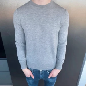 Grå stickad tröja från VNECK - Snygg grå stickad tröja från VNECK, tillverkad i Italien. Tröjan har rund halsringning, ribbade muddar och lång ärm. Perfekt för dig som gillar enkel och clean stil. Passar dig som vill ha en normal passform och ett mjukt material.