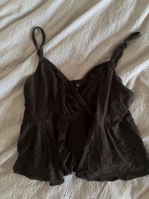 Svart omlott linnetopp - Snygg svart linnetopp med omlottdesign och smala justerbara axelband. Toppen har en lätt utsvängd passform och är perfekt för att styla med jeans eller kjol. Materialet känns mjukt och lätt mot huden. Storlek S
