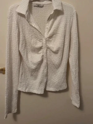 Vit strukturerad blus från Gina Tricot - Säljer en vit blus från Gina Tricot med strukturerat tyg och rynkade detaljer framtill. Blusen har krage, knappar hela vägen och långa ärmar. Materialet är mjukt och stretchigt, perfekt för en trendig look.