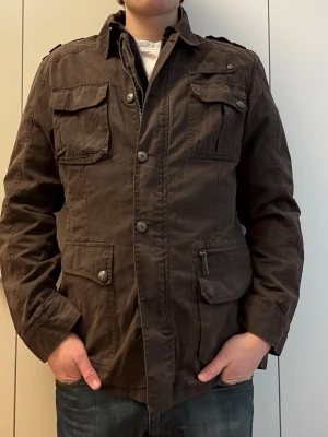 Mörkbrun utilityjacka med fickor - Snygg mörkbrun utilityjacka med fyra stora fickor framtill, axelklaffar och knappstängning. Jackan har en klassisk krage och är tillverkad i ett slitstarkt bomullsmaterial. Perfekt för dig som gillar en avslappnad men ändå stilren look.