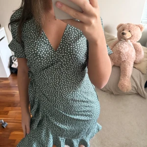 Grön prickig omlottklänning - Söt grön omlottklänning med vita prickar och volangkant nertill. Klänningen har korta ärmar, v-ringning och knyt i sidan för en snygg passform. Perfekt för sommarens alla tillfällen!