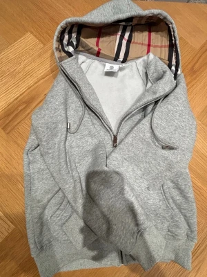 Grå zip hoodie med mönster - Snygg grå hoodie med dragkedja . Huvan har det klassiska rutmönstret i beige, rött och svart. Tröjan har ribbade muddar, fickor på sidorna och justerbara snören i huvan. Perfekt för en chill och stilren look.