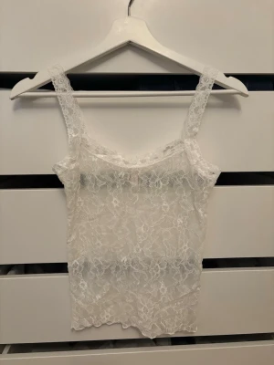 Vit spetslinne från Nelly - Säljer ett oanvänt linne i vit spets med blommigt mönster. Linnet har breda axelband och en feminin, figurnära passform. 