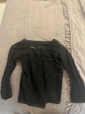 Svart virkad kofta med mönster - Snygg svart kofta med virkat mönster och lång ärm. Koftan är öppen framtill och har ett detaljerat hålmönster som ger en cool och luftig känsla. Perfekt att slänga över en topp för en trendig lager-på-lager-look.