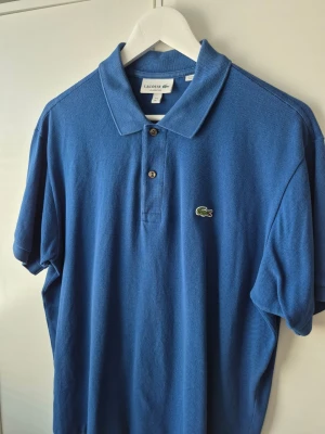Lacoste piké  - Klassisk blå pikétröja från Lacoste med broderad krokodil på bröstet. Tröjan har krage, två knappar framtill och är tillverkad i bomull. Perfekt för en stilren och sportig look.
