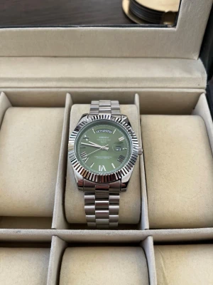 Ekstrand Daydate Grön/silver - Stilren klocka med silverfärgad länk i metall och grön urtavla. Klockan har romerska siffror, dag- och datumvisning samt räfflad bezel. Perfekt accessoar för dig som vill ha en modern och elegant look.