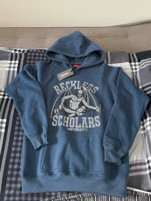 Blå hoodie Reckless Scholars - Blå hoodie från Reckless Scholars med tryck av en riddare och texten 'Reckless Scholars University' på bröstet. Hoodien har huva, känguruficka och ribbade muddar. Perfekt för en avslappnad och cool stil.