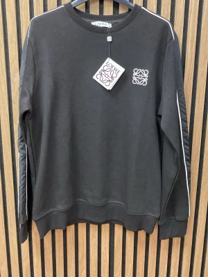 Svart sweatshirt  - Snygg svart sweatshirt  med vit broderad logga på bröstet och vita ränder längs ärmarna. Tröjan har rund halsringning, ribbade muddar och är tillverkad i mjukt bomullsmaterial. Perfekt för en stilren och avslappnad look.