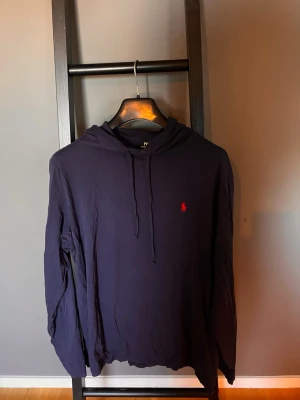  hoodie från Polo Ralph Lauren - Mörkblå hoodie med huva och dragsko från Polo Ralph Lauren. Klassisk design med röd broderad logga på bröstet. Tillverkad i mjukt och lätt material, perfekt för en avslappnad stil. Långärmad och enkel att matcha med jeans eller joggers. 