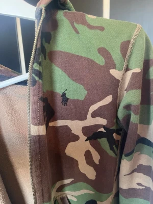 Camo hoodie från Polo Ralph Lauren - Säljer en camouflagemönstrad hoodie från Polo Ralph Lauren med dragkedja och broderad logga på bröstet. Hoodien har mjukt innerfoder och klassisk huva. Perfekt för dig som gillar streetwear och vill sticka ut lite extra.