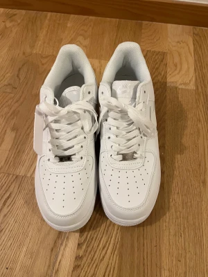 Nike Air Force 1 vita sneakers - Klassiska helvita Nike Air Force 1 sneakers i lågt utförande. Skorna har ovandel i slätt läder, perforeringar på tån för ventilation och den ikoniska swoosh-loggan på sidan. Tjock vit sula med AIR-detalj och vita skosnören. Perfekta för en clean och stilren look. Det kommer utan original förpackning 