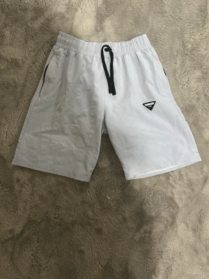 Vita shorts från Prada med snörning - Snygga vita shorts från Prada med svart snörning i midjan och det klassiska Prada-emblemet på vänster ben. Shortsen har sidofickor och en avslappnad passform, perfekta för sommardagar. Materialet känns lätt och bekvämt.